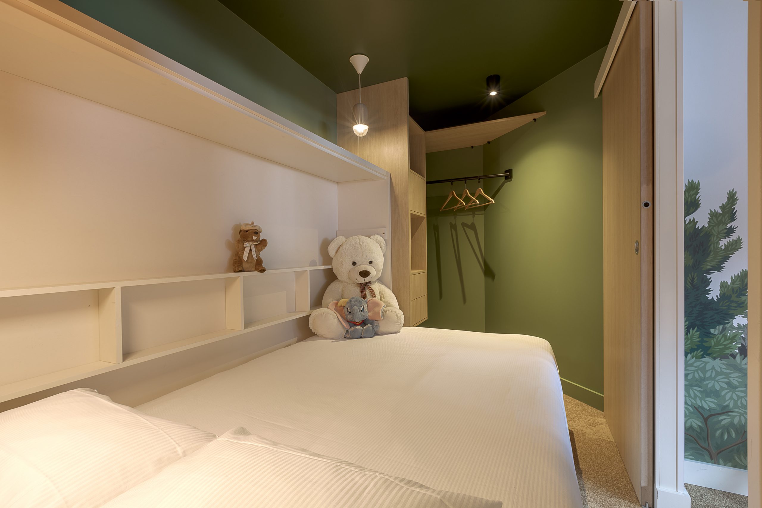 Chambre d'hôtel avec plafond vert foncé, comprenant un lit double en literie blanche et une tête de lit avec étagères blanches encastrées accueillant deux ours en peluche, un espace penderie ouvert avec tringles contre un mur vert en arrière-plan, et un papier peint mural botanique partiellement visible à travers une porte ouverte à droite, une configuration chaleureuse pour un hébergement groupe.
