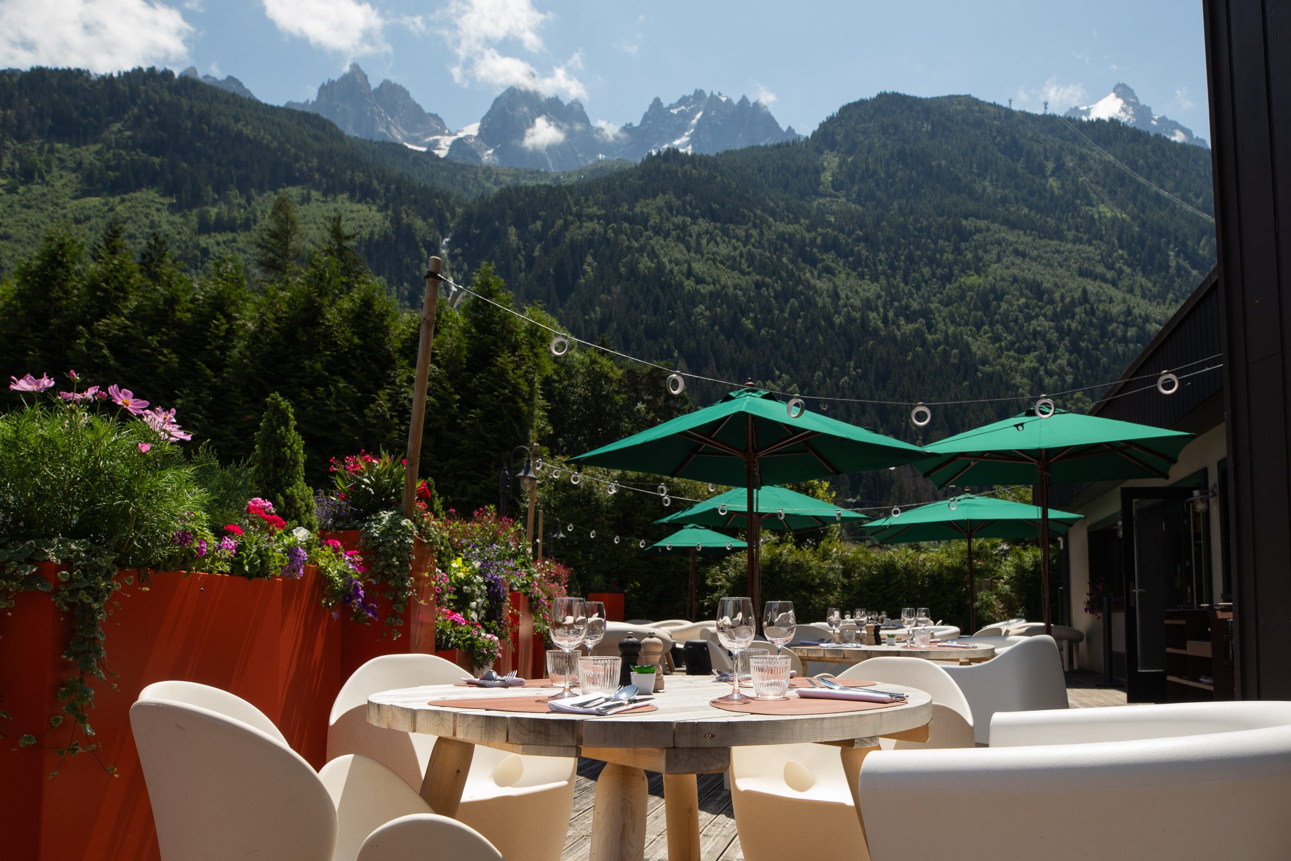 Terrasse de restaurant d'hôtel sur plancher bois, avec une table ronde dressée et chaises blanches avec verres à vin au premier plan, des parasols verts au-dessus d'autres tables au centre, des jardinières de fleurs colorées à gauche, et une vue panoramique spectaculaire sur les sommets alpins enneigés et les forêts de pins en arrière-plan, un cadre extérieur exceptionnel pour un séminaire à la montagne.