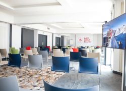 Location de salle de formation avec rangées de chaises multicolores orientées vers un grand écran à droite, dans un espace aux murs blancs lumineux et à la moquette à motif au premier plan ; l’espace de présentation et des assises supplémentaires se prolongent à l’arrière-plan.