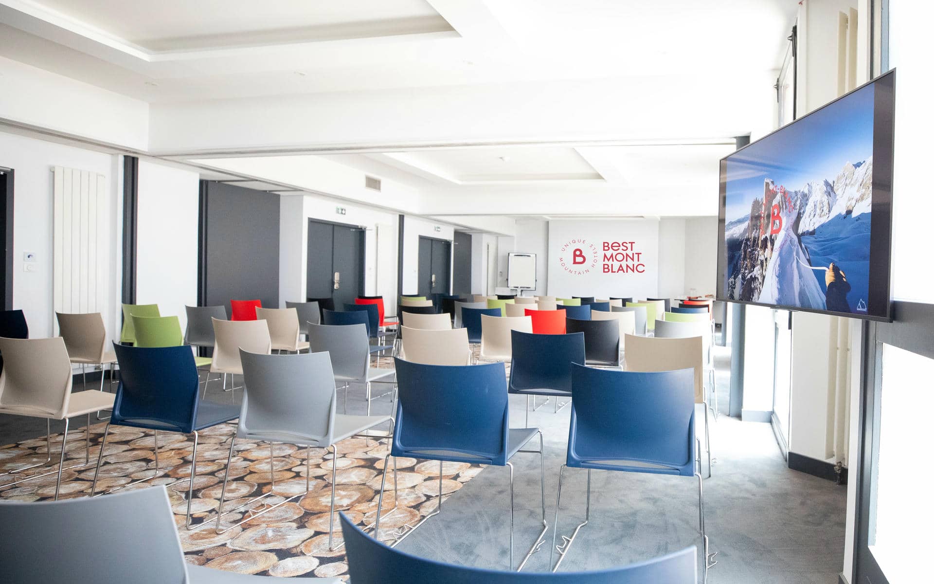 Location de salle de formation avec rangées de chaises multicolores orientées vers un grand écran à droite, dans un espace aux murs blancs lumineux et à la moquette à motif au premier plan ; l’espace de présentation et des assises supplémentaires se prolongent à l’arrière-plan.