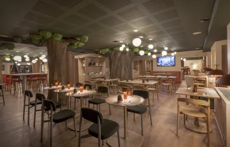 Vue générale d'un restaurant d'hôtel avec plafond vert foncé perforé orné de suspensions globes, comprenant deux grands troncs d'arbres décoratifs avec feuillage vert intégrés dans la salle, des tables rondes avec chaises sombres et bougies au centre, un espace bar avec tabourets rouges à gauche, un téléviseur mural et des banquettes rayées à droite, et un sol en bois clair, pour une restauration sur place.