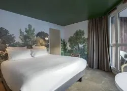 Chambre d’hôtel pour hébergement de groupe, avec au centre un grand lit double habillé de linge blanc adossé à une fresque murale représentant un paysage forestier, tandis qu’à droite une porte vitrée encadrée de longs rideaux s’ouvre sur un balcon aménagé de mobilier extérieur ; un plafond vert foncé et une moquette aux tons clairs complètent l’ensemble.