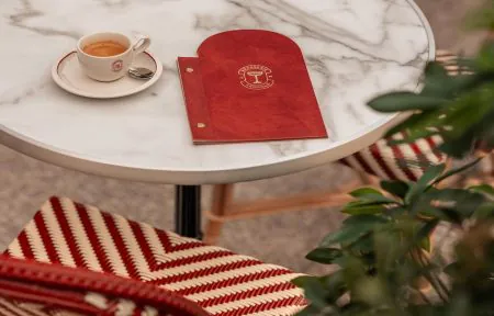 Close-up of a round white marble table with an espresso cup and saucer on the left and a red leather-bound "Brasserie Léopold" menu in the centre, surrounded by red and white striped woven bistro chairs and a green plant in the foreground on the right, on a restaurant on-site dining setting.