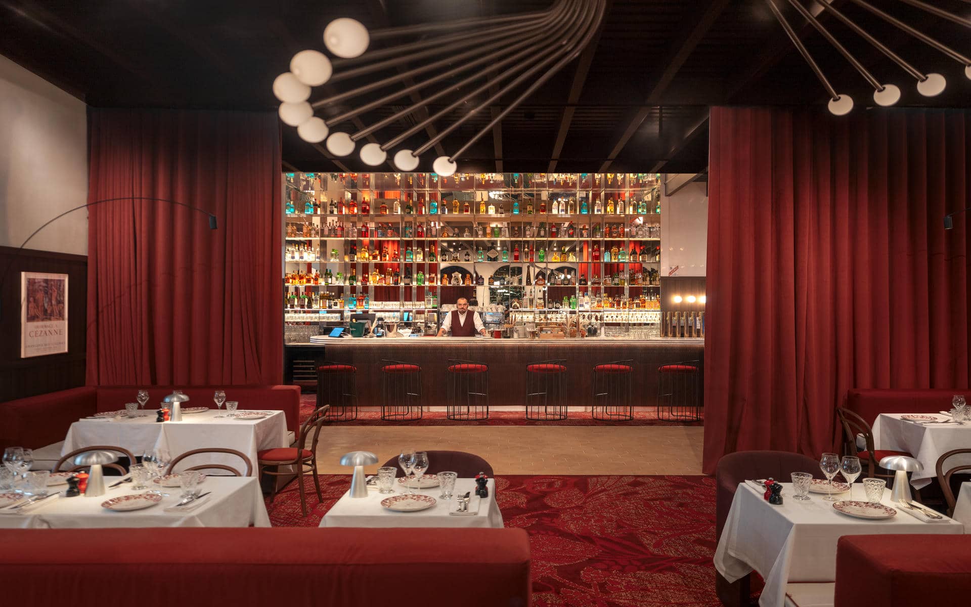 Brasserie restaurant dining room with white-clothed tables set with crystal glasses and silver lamp centrepieces, bentwood bistro chairs and dark red banquette seating on a deep red patterned carpet, large dark red curtains framing a central bar with a bartender behind a dark ribbed counter and a floor-to-ceiling backlit bottle display wall, and a sculptural multi-arm globe pendant light hanging from the dark ceiling.