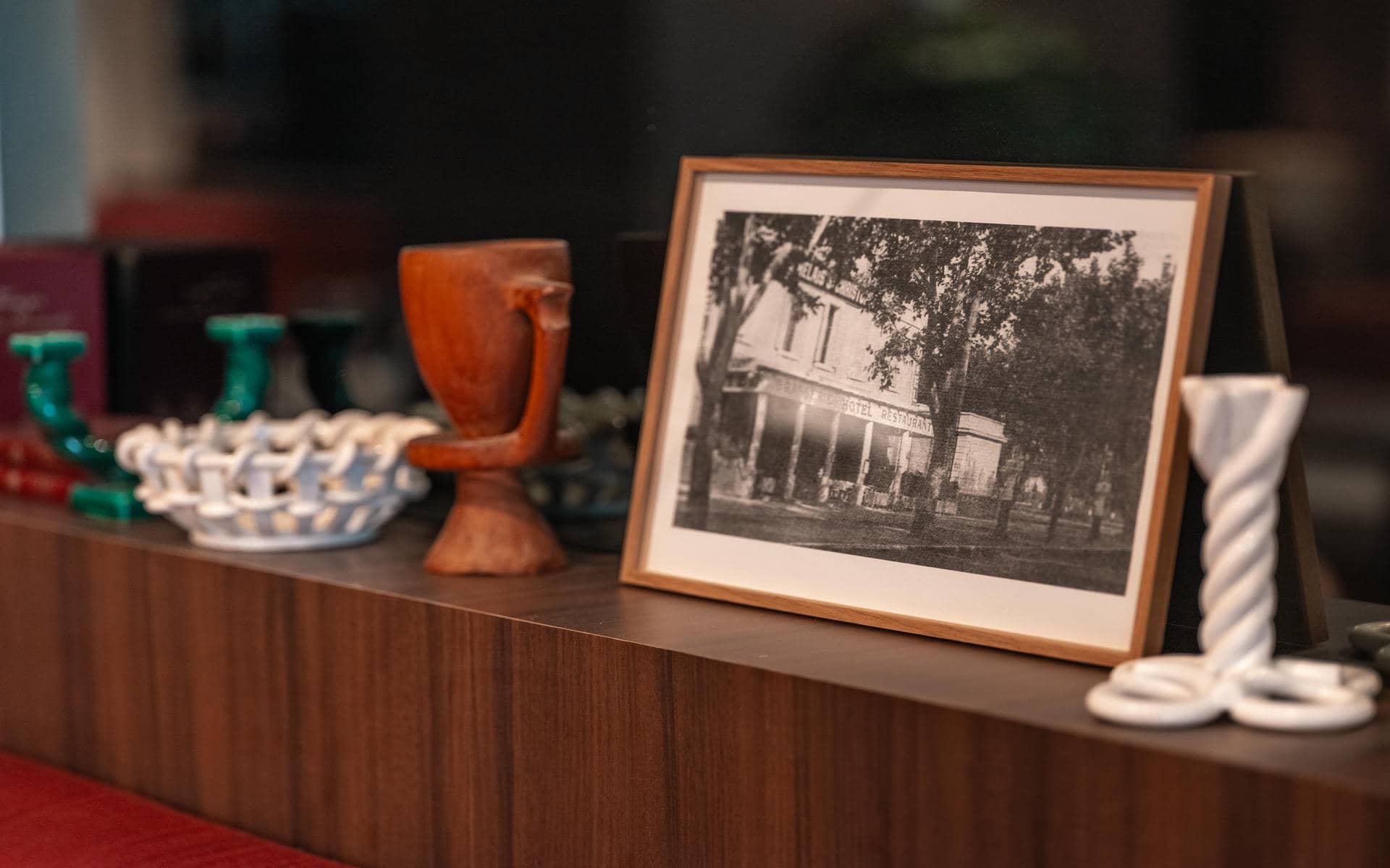 Close-up of a dark walnut wood shelf displaying decorative objects including a wood-carved goblet, a white ceramic openwork dish, two green candlesticks, a white twisted candle holder, and a framed black-and-white vintage photograph of a hotel restaurant facade at the centre, against a dark blurred background.