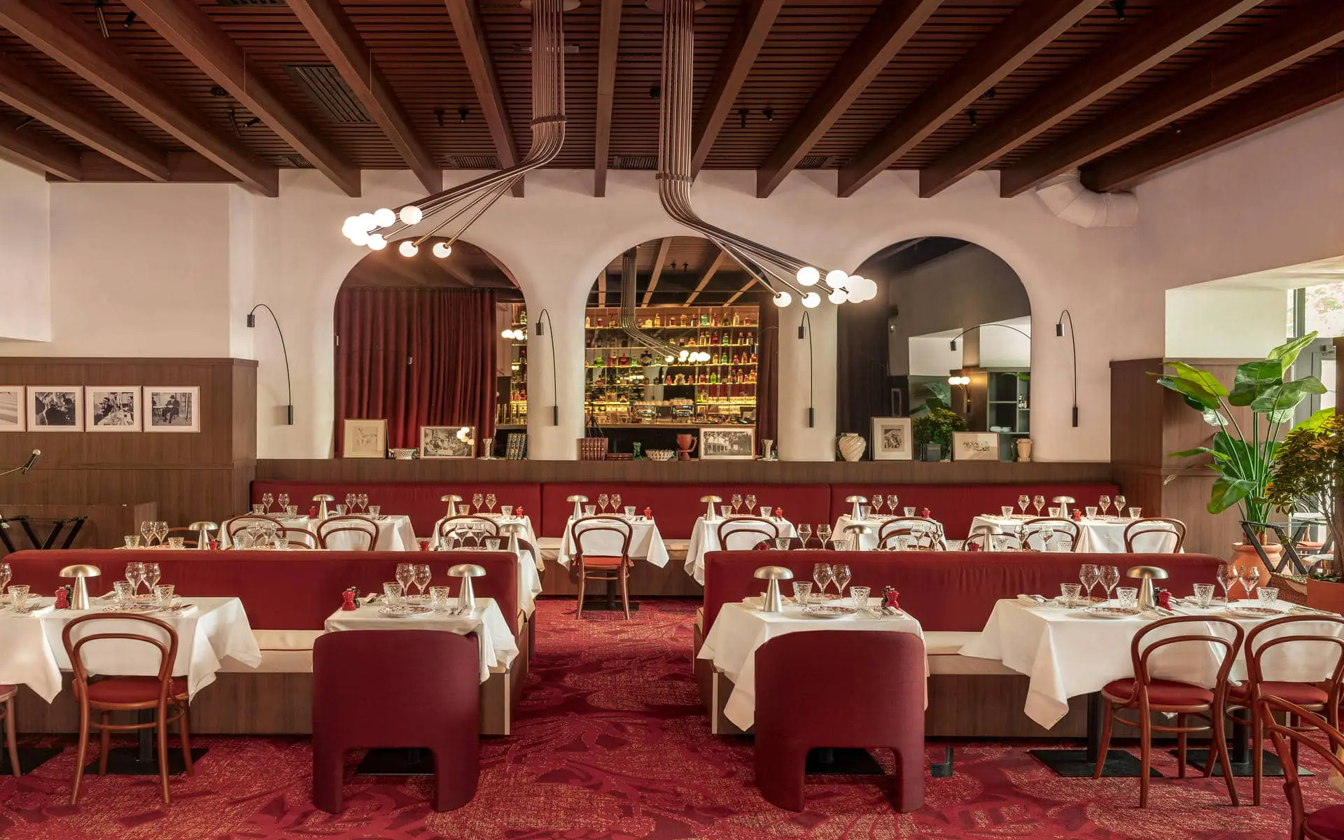 On-site dining brasserie restaurant with white-clothed tables set with crystal glasses and silver lamp centrepieces, dark red upholstered banquette seating and bentwood bistro chairs on a deep red patterned carpet, two arched openings revealing a backlit bottle display bar in the background, dark wood beam ceiling with globe pendant lights, black-and-white framed photographs on the left wall, and a large green plant on the right.