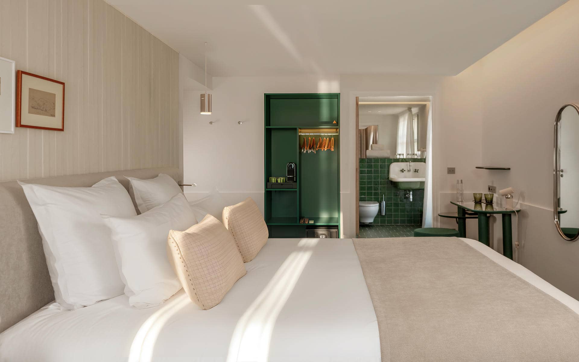 Hotel group accommodation bedroom with a double bed dressed in white linen and beige woven cushions against a light wood-panelled headboard wall, a dark green open wardrobe unit with lit hanging rail and coffee machine in the centre background, an open doorway revealing a green-tiled bathroom with a vintage white wall-mounted sink on the right, a small green-legged glass console table, and a chrome oval floor mirror on the far right.