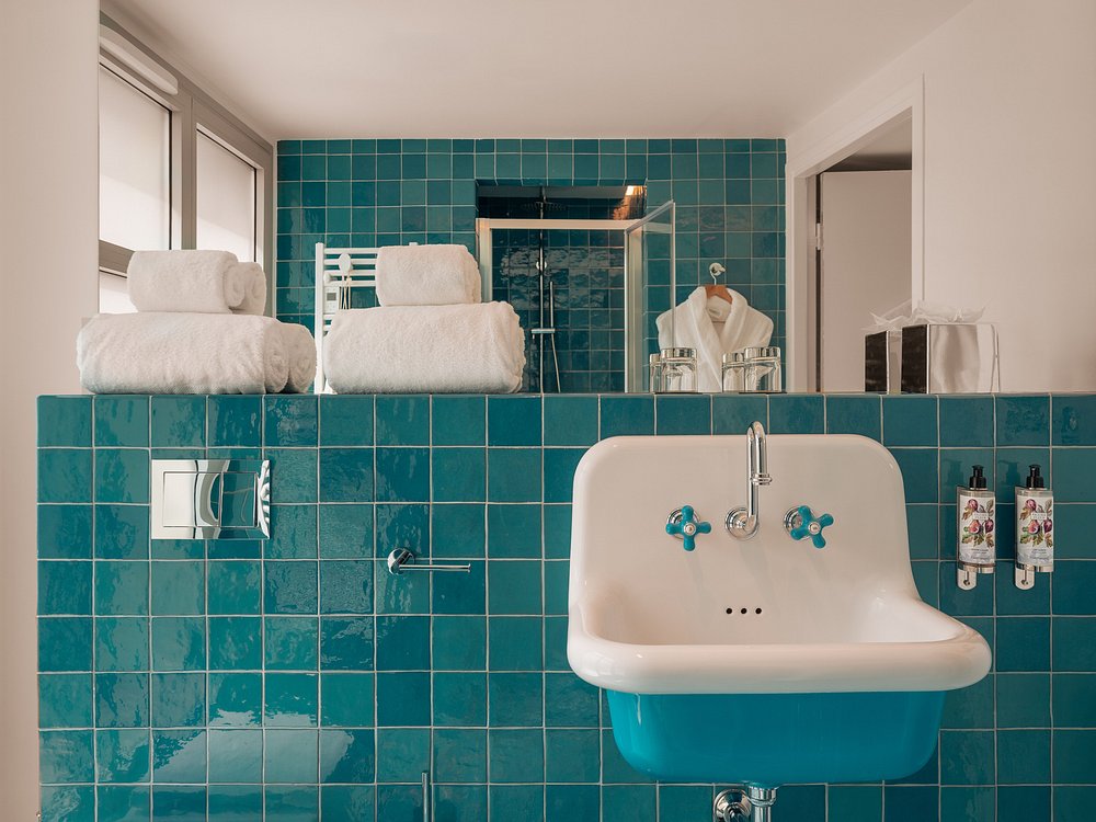 Salle de bain de chambre d'hébergement groupe avec des carreaux carrés bleu canard brillants couvrant les murs, un lavabo vintage blanc mural à poignées de robinetterie en céramique bleu canard et accessoires chromés au premier plan, deux serviettes blanches pliées et des distributeurs de produits de toilette en verre sur l'étagère carrelée au-dessus, un grand miroir reflétant une cabine de douche vitrée et un peignoir blanc sur un cintre, et deux distributeurs de produits muraux à droite.
