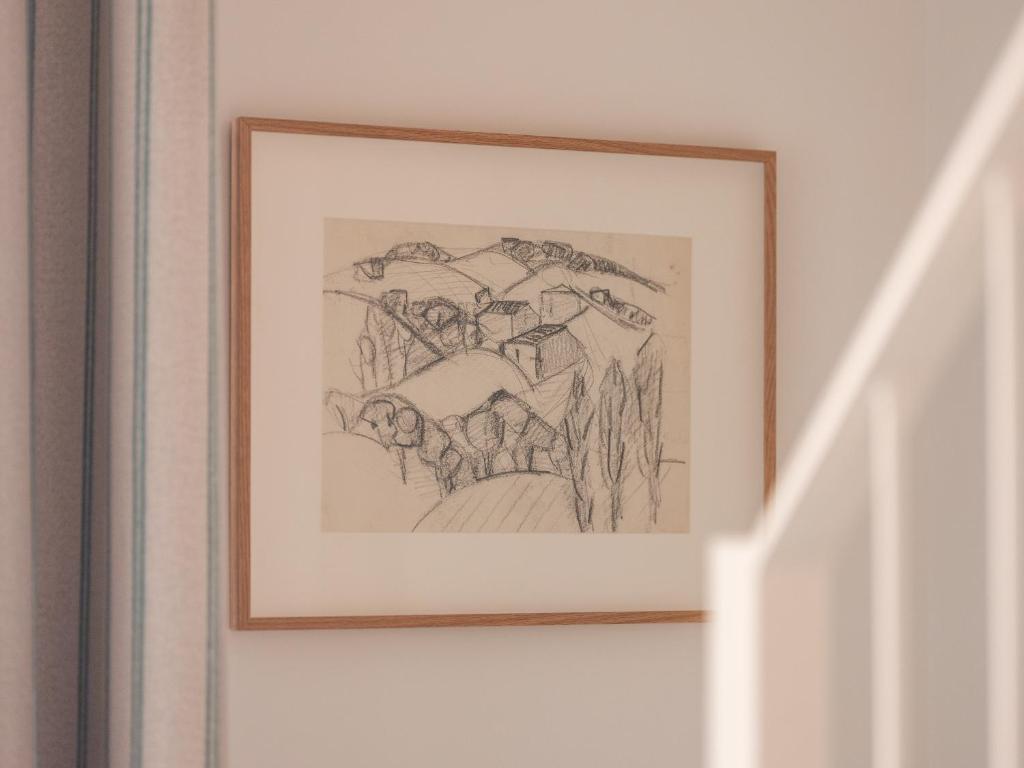 Close-up of a framed pencil sketch hung on a white wall, depicting a Provençal landscape with rolling hills, cypress trees and stone village houses, set in a thin natural wood frame, with a striped curtain visible on the left edge.