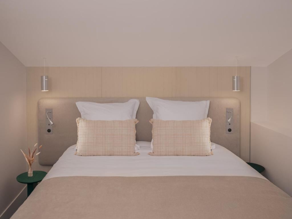 Close-up of a hotel double bed with a beige bedspread, white pillows and two textured blush cushions against a light wood headboard wall with silver reading sconces on each side and small dark green bedside tables, offering comfortable group accommodation.