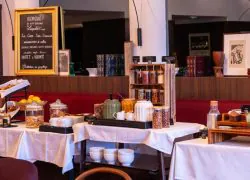 Hotel breakfast buffet spread across white-clothed tables in a warm-lit room with wood panelling and a red banquette, featuring baskets of bread, glass jars of cereals and cookies, a kettle, bowls and assorted condiments, with a chalkboard welcome sign on the left and framed artwork in the background, everything you need to start a study day on the right foot.