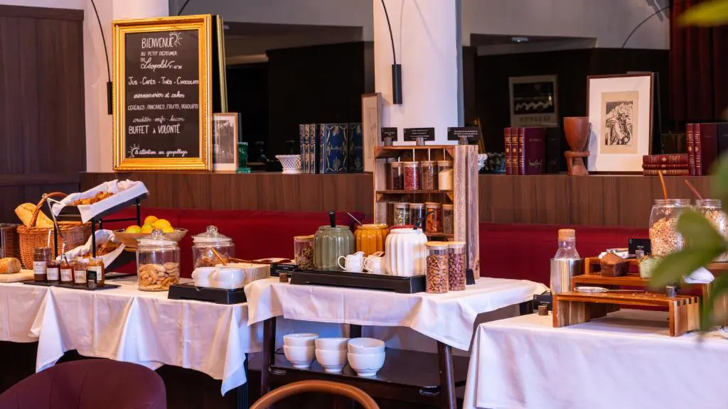 Hotel breakfast buffet spread across white-clothed tables in a warm-lit room with wood panelling and a red banquette, featuring baskets of bread, glass jars of cereals and cookies, a kettle, bowls and assorted condiments, with a chalkboard welcome sign on the left and framed artwork in the background, everything you need to start a study day on the right foot.