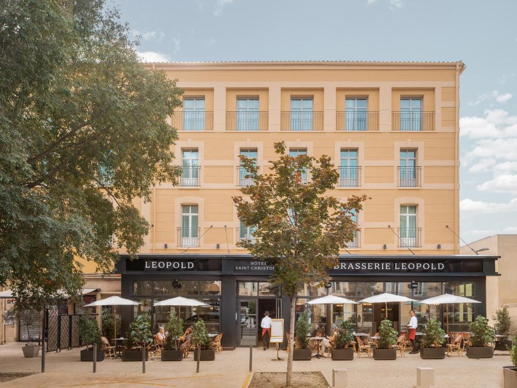 Exterior facade of the Leopold hotel and brasserie, a four-storey ochre-yellow building with wrought-iron balconies, featuring a dark shopfront with white signage; the terrace in the foreground is set with rattan chairs, white parasols and potted plants, ideal for an urban seminar venue.