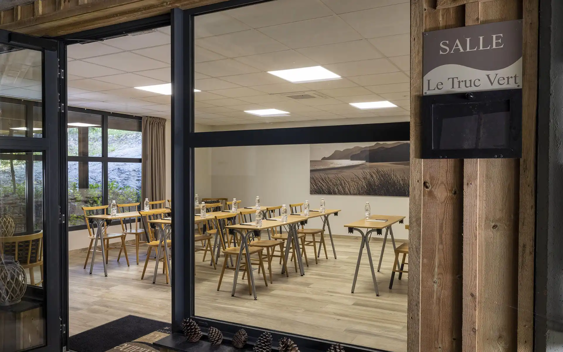 Classroom-style meeting room with light wood flooring, featuring rows of tables and wooden chairs arranged facing the front across the center and background, large glazed openings on the left, and a landscape photograph mounted on the rear wall; bright ceiling lighting and neutral-toned finishes create a clean, professional setting.