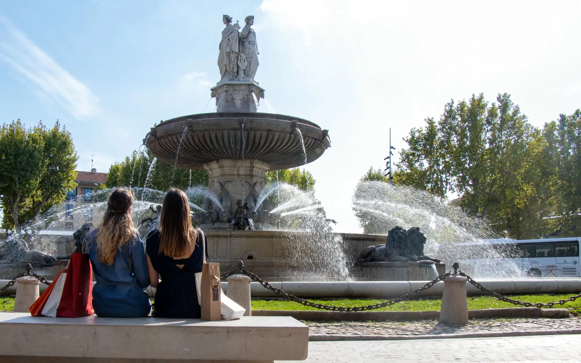 Monumental fountain in Aix-en-Provence, featuring a large circular basin crowned by statues at the center, powerful water jets and sculpted lions in the foreground, while two seated visitors observe the scene from a stone bench on the left.