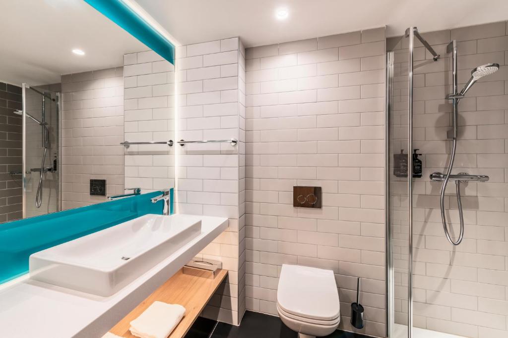 Hotel bathroom with white subway tiles and dark floor, featuring a rectangular vessel sink on a wooden shelf with a turquoise-framed mirror on the left, a wall-hung toilet in the centre, and a glass-enclosed shower with a chrome thermostatic column on the right.
