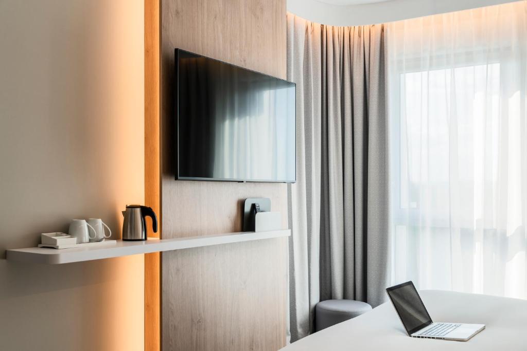 Hotel room detail with a light wood panel featuring a wall-mounted television and a white shelf with a kettle and cups, warm LED lighting on the left, and a large window with grey curtains and a laptop on a desk in the background.