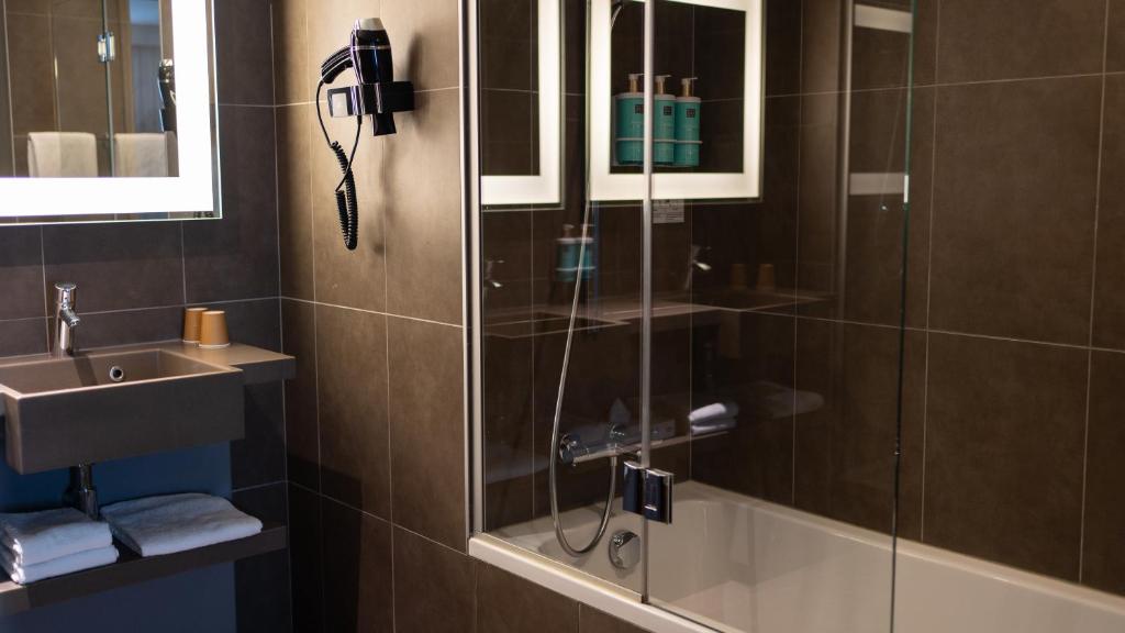 Salle de bain d'hôtel adaptée pour un séminaire en ville, comprenant un lavabo en pierre grise avec serviettes pliées et miroir lumineux à gauche, un sèche-cheveux noir fixé au mur carrelé marron, et une baignoire vitrée avec mitigeur chromé et flacons de soin bleu canard reflétés dans le miroir à droite.
