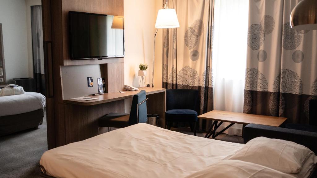 Chambre d'hôtel adaptée pour un hébergement groupe, comprenant un lit beige au premier plan, un bureau en bois avec télévision à écran plat, téléphone et chaise bleue au centre, un petit fauteuil rond et une table basse en bois à droite, et un second lit visible à travers une cloison ouverte à gauche.