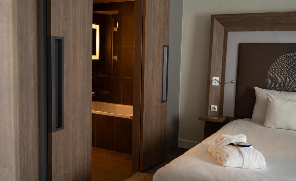 Chambre d'hôtel adaptée pour un hôtel pour séminaire, comprenant un lit blanc avec peignoir plié et masque de nuit au premier plan, une tête de lit en bois avec liseuse à droite, et des portes coulissantes en bois s'ouvrant sur une salle de bain carrelée en marron avec baignoire et miroir lumineux en arrière-plan.