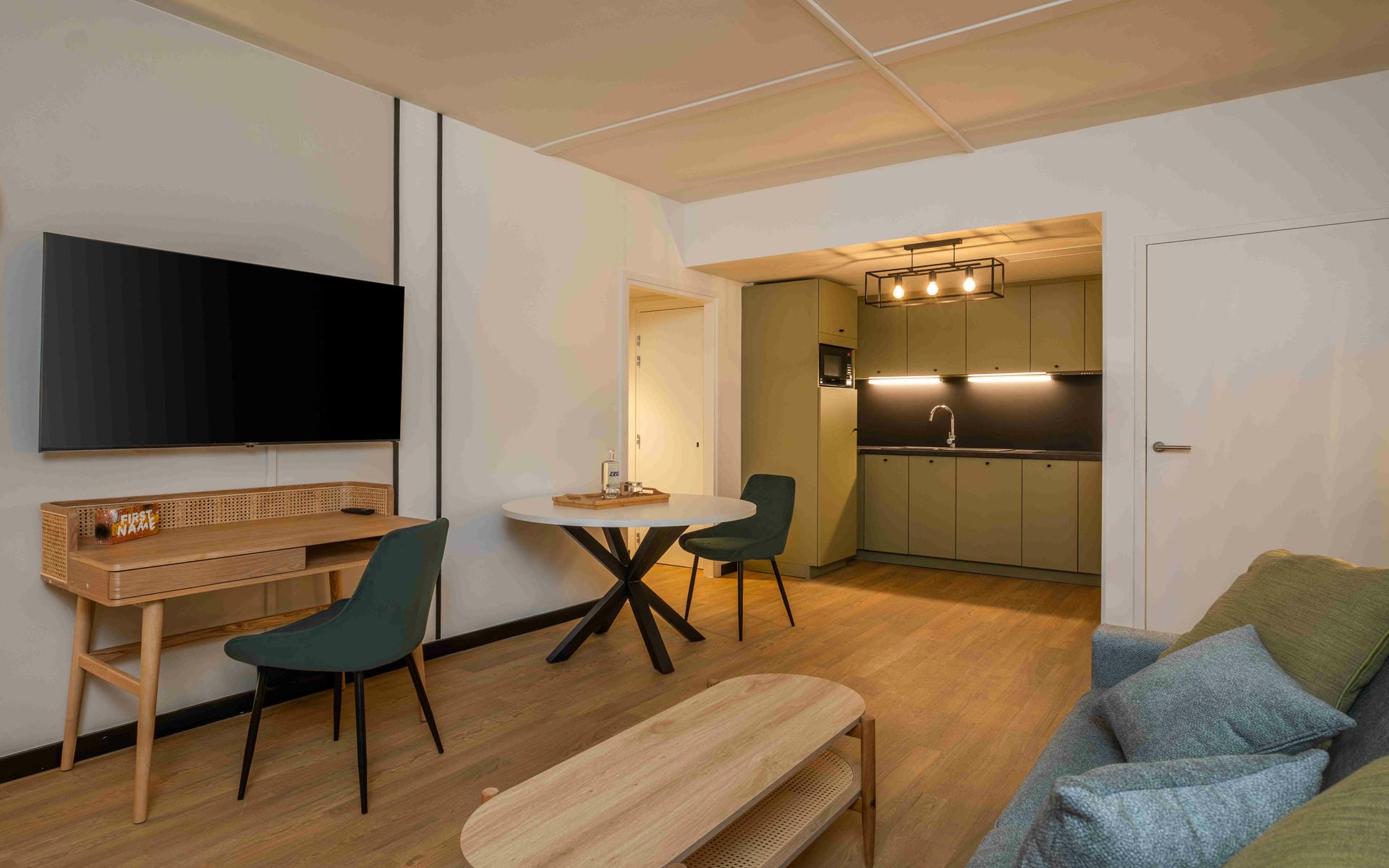 Hotel suite living area with wooden flooring, featuring a wall-mounted television and desk on the left, a round dining table at the center, and an open kitchenette with olive-green cabinetry in the background; a sofa and wooden coffee table occupy the foreground on the right.