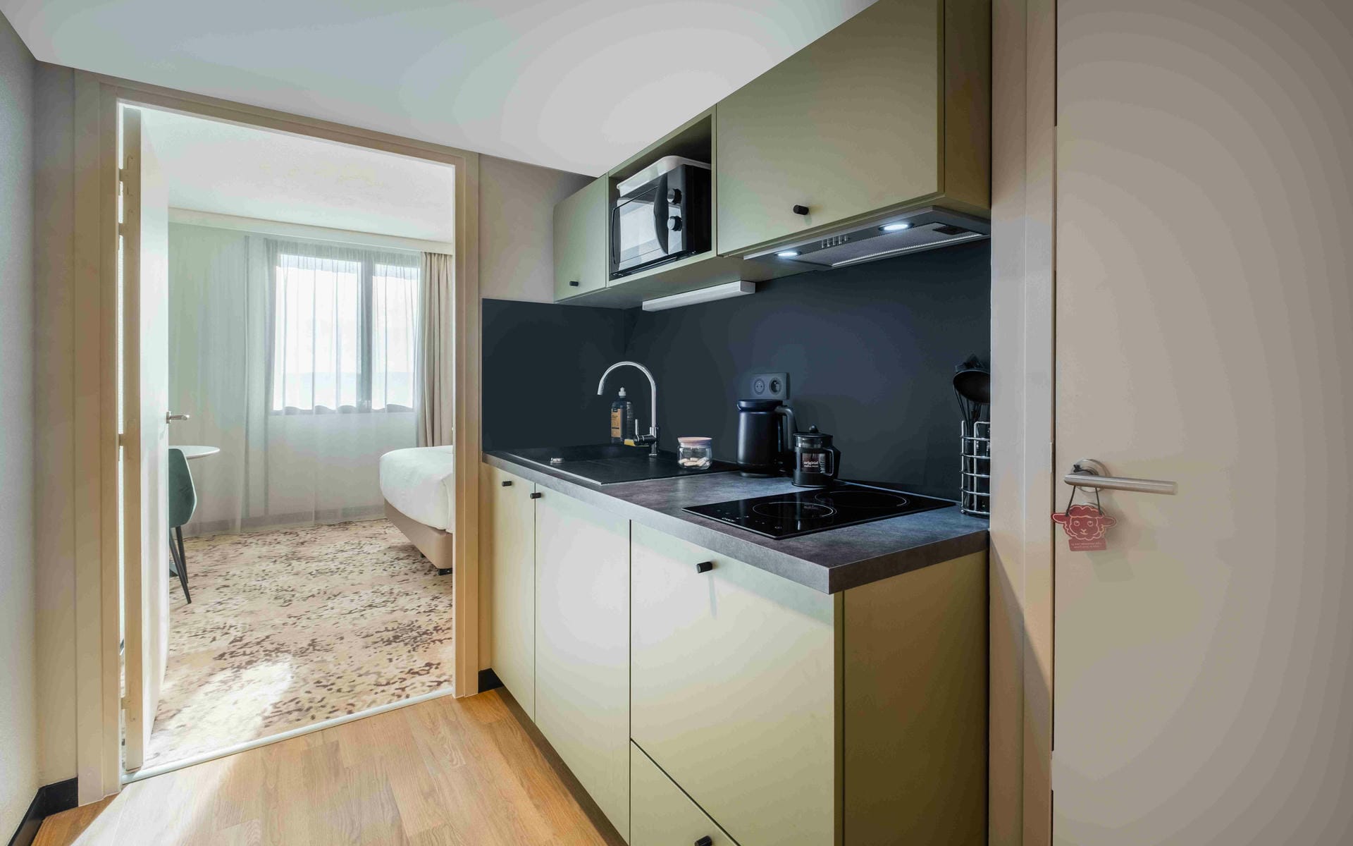 Hotel suite kitchenette with light wood cabinetry, a dark countertop, sink, cooktop and microwave on the right; an open doorway leads to a bright bedroom with patterned carpet, a bed and a small table near the window.