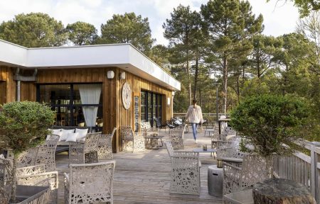 Outdoor bar terrace with wooden decking, featuring patterned light-colored lounge chairs and low tables arranged across the foreground, a wood-clad building with large windows on the left, and a wooded landscape in the background; a guest walks along the central pathway between seating areas.
