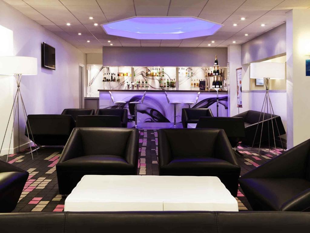 Bar lounge d'hôtel avec moquette foncée à motifs, fauteuils en cuir noir disposés autour d'une table basse blanche au premier plan, deux lampadaires blancs de part et d'autre, et un comptoir de bar rétroéclairé avec bouteilles sur étagères et tabourets blancs sous un plafond alcôve éclairé en violet en arrière-plan, un cadre idéal pour un afterwork.