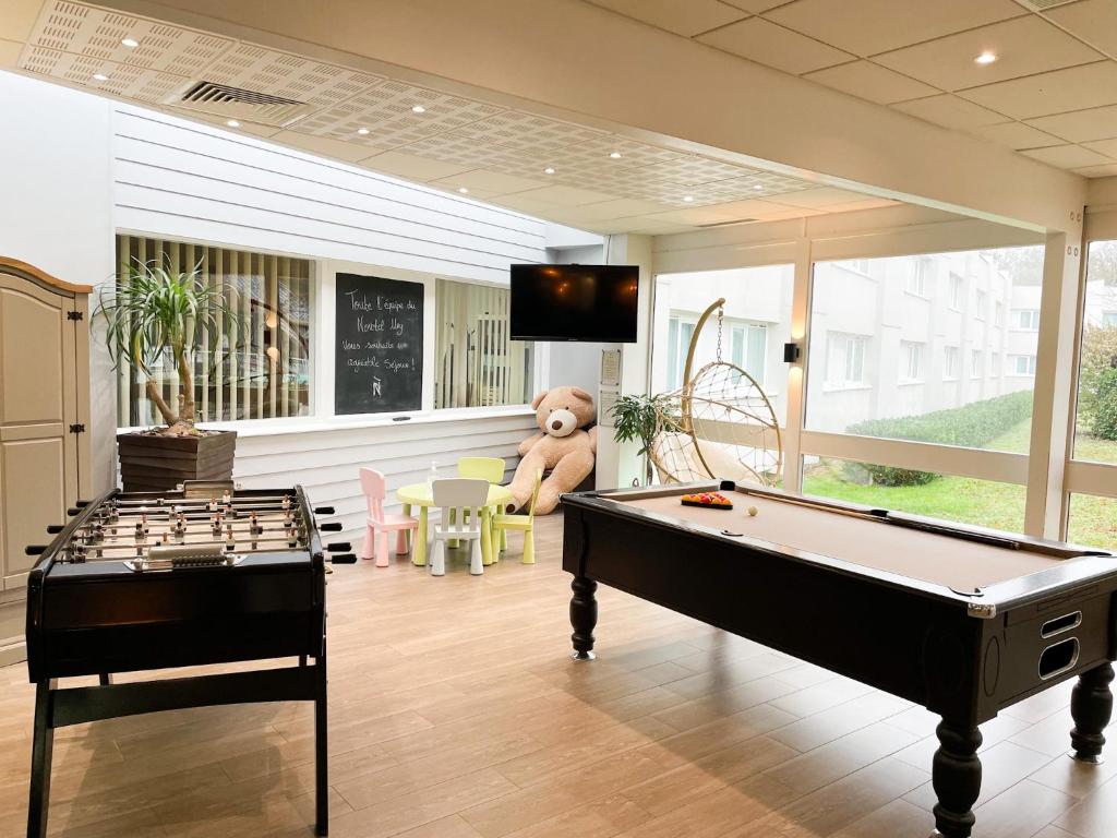 Salle de jeux intérieure avec sol en bois clair, un baby-foot noir à gauche et un billard marron à droite au premier plan, une petite table d'enfants avec chaises pastel, un grand ours en peluche et un fauteuil suspendu en rotin près d'une télévision murale au plan intermédiaire, et de grandes fenêtres donnant sur un bâtiment et un jardin en arrière-plan, un espace de détente au sein de cet hébergement groupe.