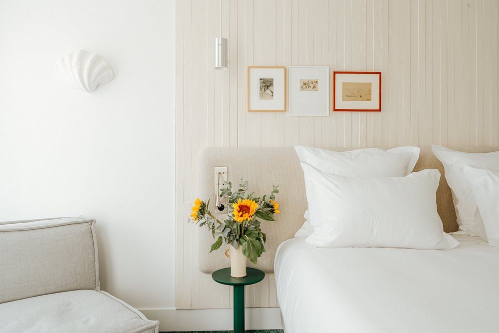 Hotel group accommodation bedroom detail with a double bed dressed in white linen against a light wood-panelled headboard wall, three small framed artworks above, a small dark green round bedside table with a white vase of sunflowers and eucalyptus, a chrome reading sconce, a white decorative shell wall ornament on the left wall, and the arm of a light grey sofa visible in the foreground on the left.