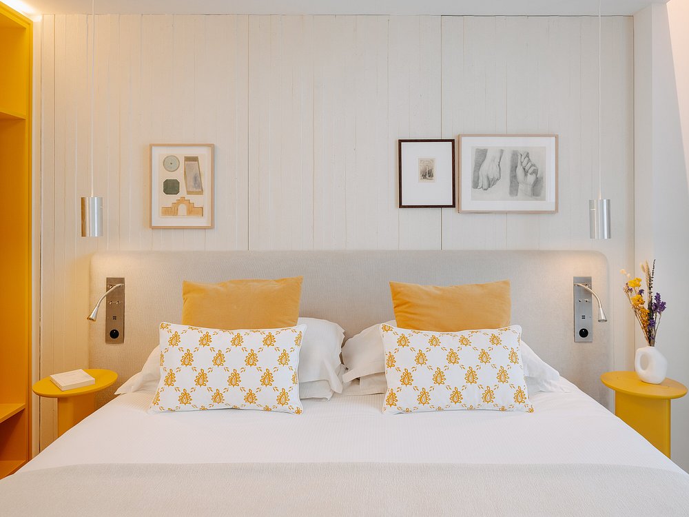 Hotel group accommodation bedroom with a double bed dressed in white linen with yellow cushions and two white patterned yellow floral cushions, a light grey upholstered headboard against a white wood-panelled wall with three framed artworks above, two chrome reading sconces and yellow round bedside tables on each side, a white ceramic vase with dried yellow and purple flowers on the right, and a yellow open wardrobe unit visible on the left.