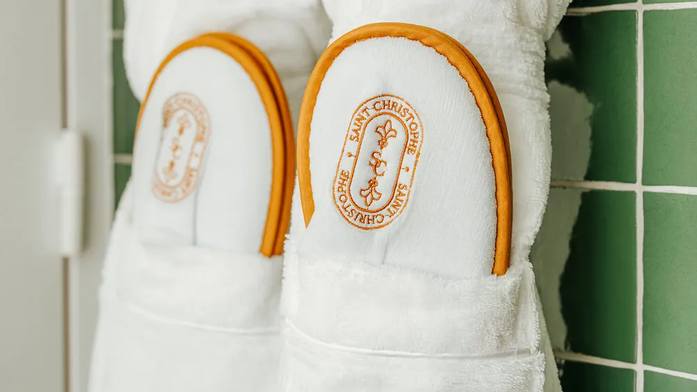Close-up of two pairs of white hotel slippers with orange piping and an embroidered "Saint-Christophe" oval logo, tucked into the breast pockets of white terry cloth bathrobes with orange piping, against a green square-tiled bathroom wall.
