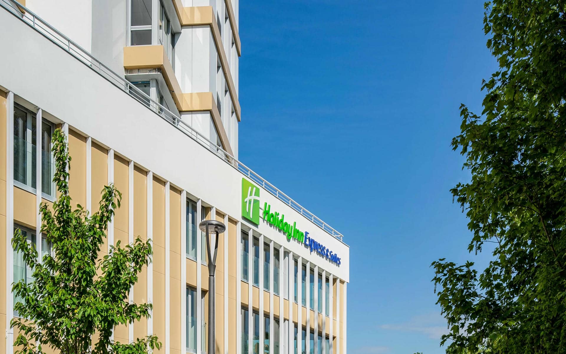 Exterior facade of the Holiday Inn Express & Suites hotel for a city seminar, with a beige and white multi-storey building featuring large vertical windows, a green and blue illuminated brand sign at the centre, a street lamp in the foreground, green trees on both sides, and a clear blue sky in the background.