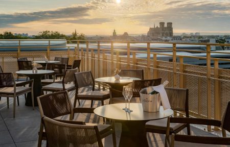 Terrasse afterwork sur rooftop urbain avec tables rondes gris clair et chaises en corde tressée sombre sur sol carrelé, un seau à champagne avec deux flûtes au premier plan, une rambarde en métal perforé doré, et une vue panoramique sur le coucher de soleil au-dessus de la skyline de Reims avec la cathédrale en silhouette en arrière-plan.