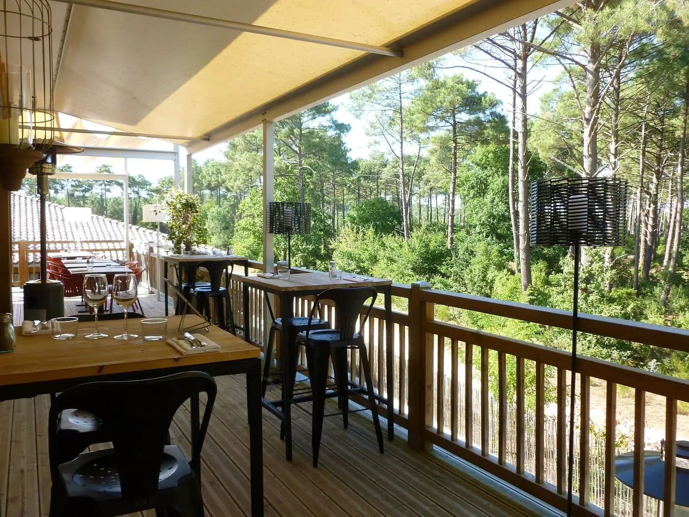 Shaded restaurant terrace with wooden decking, featuring dining tables and high-top tables with black stools along the foreground and center, a wooden railing on the right, and a panoramic view of a pine forest in the background; a retractable awning overhead provides shelter and filtered light.