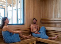 en::Large wooden sauna with views of the surroundings, with two people in blue towels, in a Chamonix seminar hotel. fr::Grand sauna en bois avec vue sur les environs, deux personnes en serviettes bleues, dans un hôtel séminaire à Chamonix.