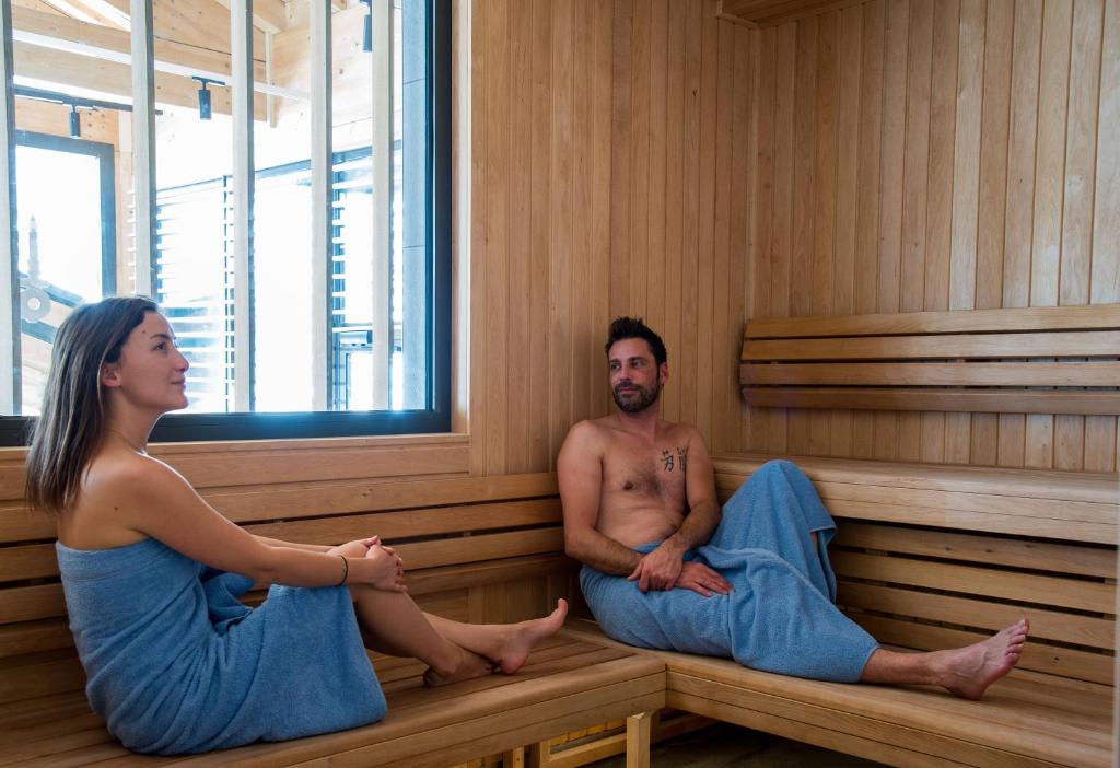 en::Large wooden sauna with views of the surroundings, with two people in blue towels, in a Chamonix seminar hotel. fr::Grand sauna en bois avec vue sur les environs, deux personnes en serviettes bleues, dans un hôtel séminaire à Chamonix.
