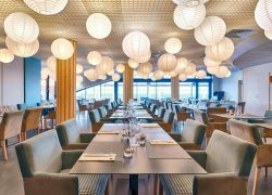 Salle de restaurant d'hôtel avec des dizaines de suspensions rondes en papier blanc accrochées à un plafond à lattes en bois, comprenant des tables sombres dressées avec verres à vin et serviettes pliées au premier plan, des fauteuils en velours bleu canard et beige disposés le long de tables à structure bois au centre, et de grandes baies vitrées laissant apparaître une vue sur la mer en arrière-plan sous une lumière naturelle, idéal pour la restauration sur place.