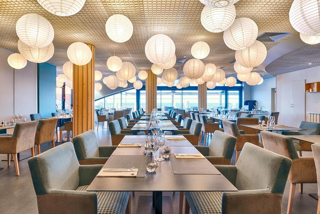 Salle de restaurant d'hôtel avec des dizaines de suspensions rondes en papier blanc accrochées à un plafond à lattes en bois, comprenant des tables sombres dressées avec verres à vin et serviettes pliées au premier plan, des fauteuils en velours bleu canard et beige disposés le long de tables à structure bois au centre, et de grandes baies vitrées laissant apparaître une vue sur la mer en arrière-plan sous une lumière naturelle, idéal pour la restauration sur place.