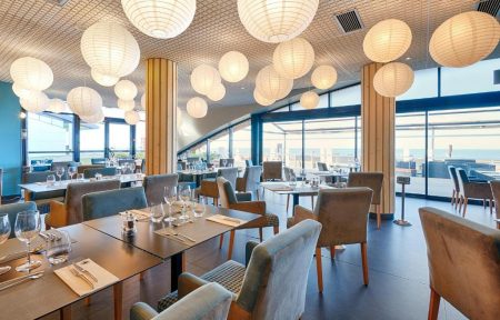 Restaurant sur place d'hôtel avec des tables en bois foncé dressées de verres à vin, serviettes et couverts, encadrées de chaises rembourrées grises et bleues au premier plan, des colonnes en bois et d'autres tables dressées au centre, et des baies vitrées ouvrant sur une vue mer sous de nombreuses suspensions en papier rondes en arrière-plan.