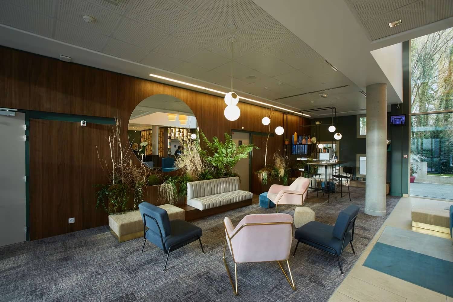 Hotel lobby lounge ideal for a city seminar, with pink and dark blue armchairs arranged around a low table in the foreground, a beige upholstered bench and green plants beside a round mirror against a wood-panelled wall, hanging spherical pendant lights above, a concrete column and glazed entrance opening onto trees on the right, and grey patterned carpet flooring.