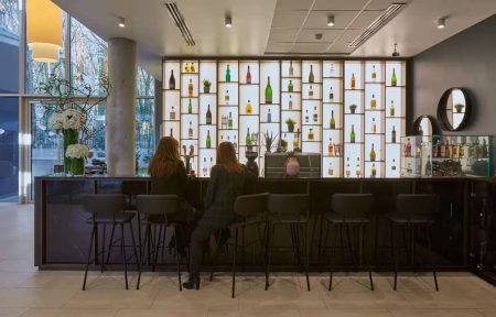 Hotel bar area for after-work drinks with a dark wooden counter and black bar stools in the foreground, two women seated at the bar facing an illuminated backlit shelving wall displaying rows of bottles, round mirrors and a large white floral bouquet on the left, and beige floor tiles with tall windows revealing trees outside in the background.