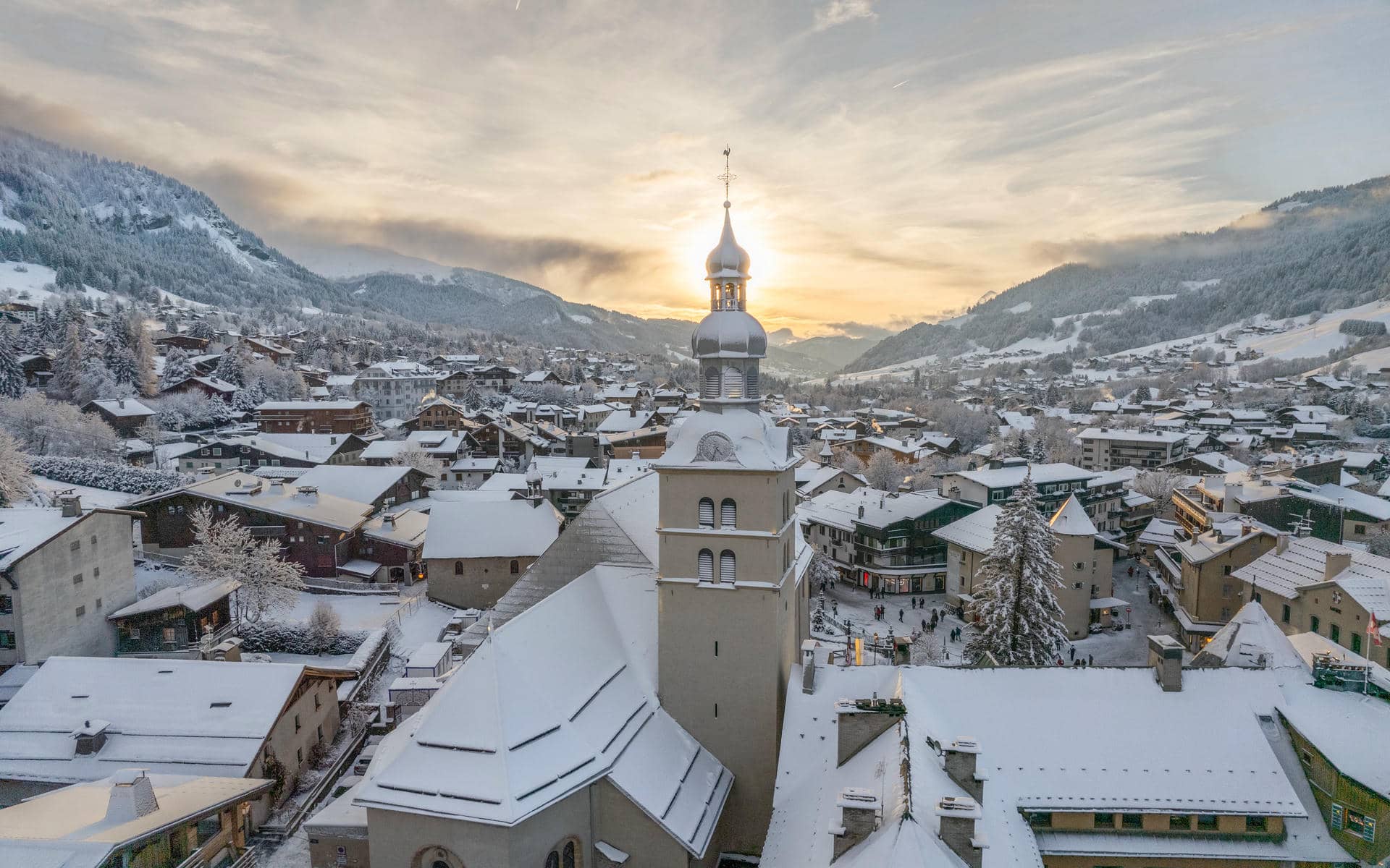 Vue aérienne d'ensemble de Megève, destination de séminaire à la montagne, montrant le clocher à bulbe de l'église du village alpin enneigé rétroéclairé par le soleil couchant au centre, des toits de chalets et de petites rues où circulent des passants en contrebas, et des montagnes enneigées boisées s'élevant de part et d'autre sous un ciel doré et voilé.