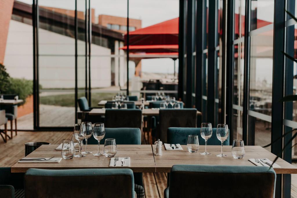 Restaurant sur place d'hôtel avec une table en bois dressée de verres à vin, serviettes et couverts, encadrée de chaises en velours bleu canard au premier plan, d'autres tables dressées avec des chaises assorties au centre, et des baies vitrées révélant une terrasse avec parasols rouges et des bâtiments en arrière-plan.