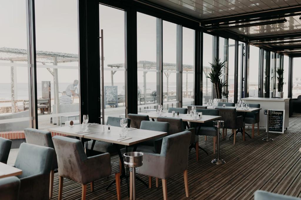 Restaurant sur place d'hôtel avec des tables en bois dressées de verres à vin et des fauteuils rembourrés gris sur pieds en bois clair au premier plan, un seau à glace sur pied métallique et une ardoise menu au centre, des plantes vertes en pot près d'un comptoir de service blanc à droite, et des baies vitrées à cadres noirs du sol au plafond donnant sur une terrasse en bois et une vue sur la mer en arrière-plan.