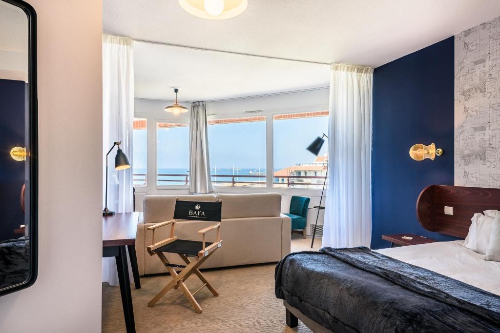 Chambre d'hôtel avec lit et mur d'accent bleu foncé à tête de lit en bois incurvée à droite, canapé beige avec chaise pliante de metteur en scène à l'enseigne de l'hôtel et bureau en bois à gauche, fauteuil bleu canard près de baies vitrées du sol au plafond donnant sur la mer et une ville côtière en arrière-plan, et grand miroir appuyé contre le mur à gauche, une chambre adaptée à un séminaire en bord de mer.