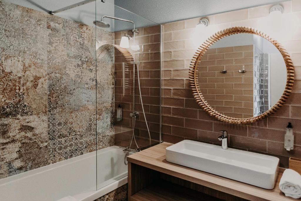 Salle de bain d'hôtel avec baignoire blanche équipée d'un pare-douche vitré et d'une douche de tête à effet pluie à gauche contre un mur carrelé à motif mosaïque, et meuble-vasque en bois avec vasque rectangulaire blanche sous un miroir rond à cadre en rotin sur carrelage effet brique brun à droite, adaptée à un séminaire en bord de mer.