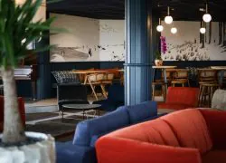 Espace bar-salon d'hôtel avec une grande plante verte au premier plan, des canapés en velours orange et bleu marine autour d'une table basse ronde en métal noir au centre, des tabourets de bar en bois et des chaises en rotin près de tables hautes en arrière-plan, des colonnes bleu foncé et une fresque murale illustrée en noir et blanc représentant un paysage côtier, avec des suspensions globe lumineuses au-dessus, parfait pour un afterwork.