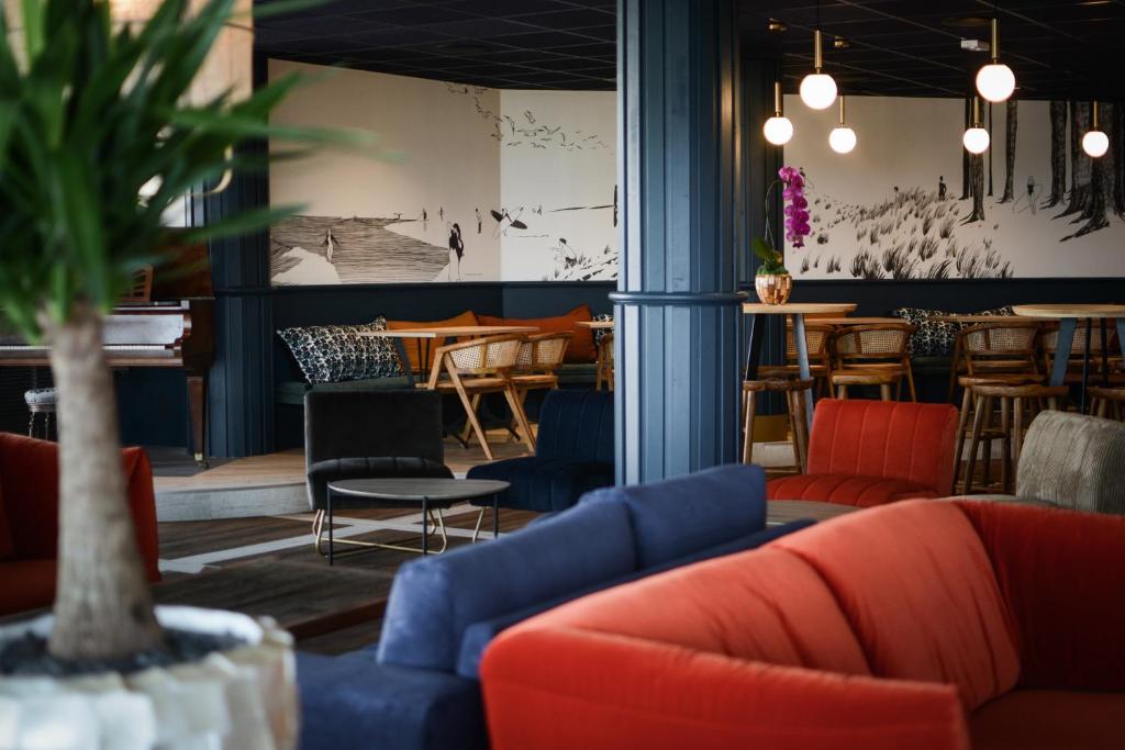 Espace bar-salon d'hôtel avec une grande plante verte au premier plan, des canapés en velours orange et bleu marine autour d'une table basse ronde en métal noir au centre, des tabourets de bar en bois et des chaises en rotin près de tables hautes en arrière-plan, des colonnes bleu foncé et une fresque murale illustrée en noir et blanc représentant un paysage côtier, avec des suspensions globe lumineuses au-dessus, parfait pour un afterwork.