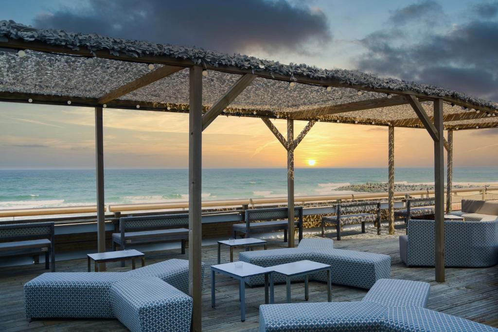 Terrasse afterwork en bord de mer avec des assises modulables grises à motifs et de petites tables métalliques au premier plan, un espace en caillebotis bois avec bancs sous une pergola en bois recouverte de chaume au centre, et la mer avec des vagues visibles sous un ciel de coucher de soleil en arrière-plan.